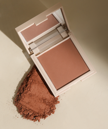 An open square makeup compact with a mirror on the lid and a pressed powder pan in a warm medium tan-brown shade, shown on a clean white background.